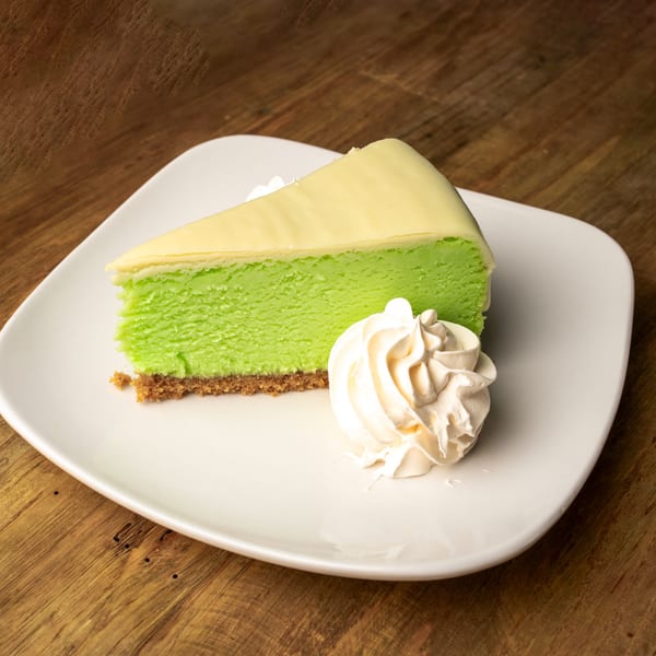 A slice of vibrant green cheesecake with a graham cracker crust and whipped cream on a white plate.