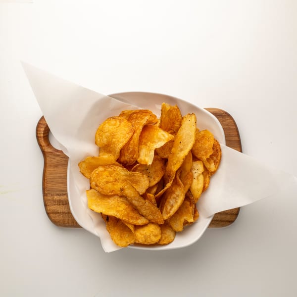 A white bowl filled with golden-brown, seasoned potato chips, lined with white parchment paper, on a wooden board.