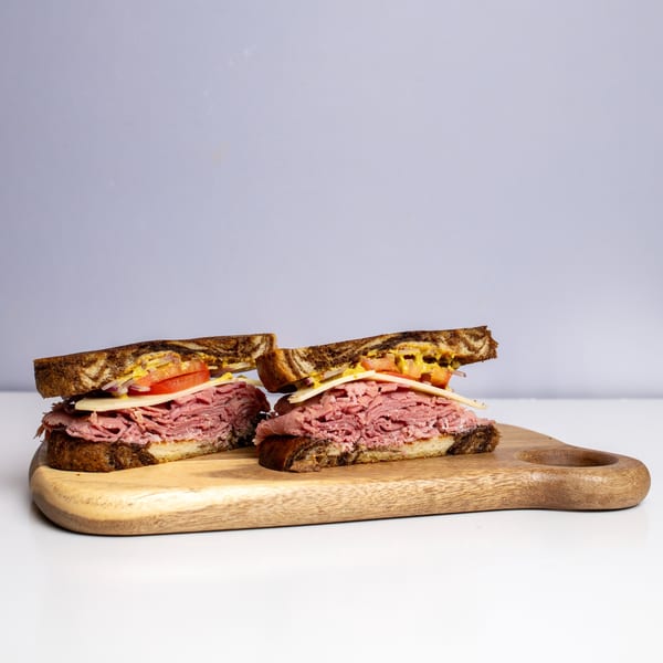 Two halves of a deli sandwich with pastrami, cheese, tomato, and mustard on rye bread.