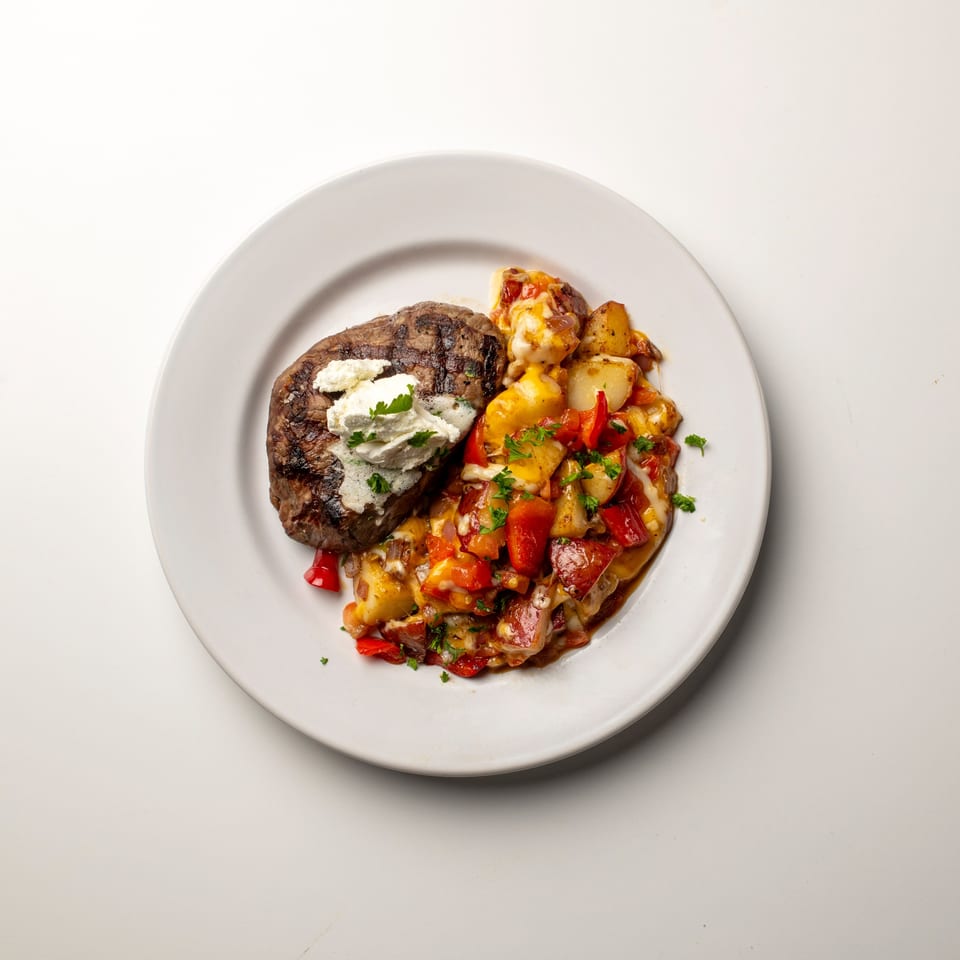 Grilled steak topped with white cheese and parsley, served with roasted potatoes and red peppers.