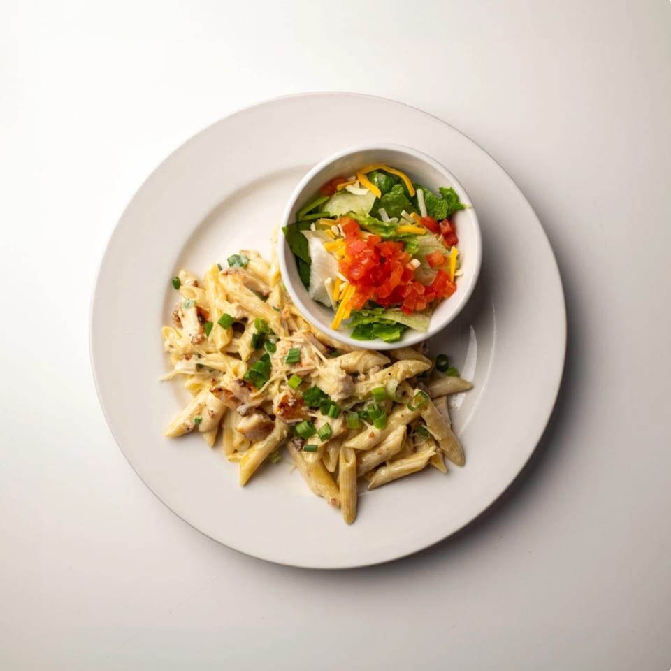 Creamy penne pasta with chicken and green onions, served with a side salad in a white bowl.