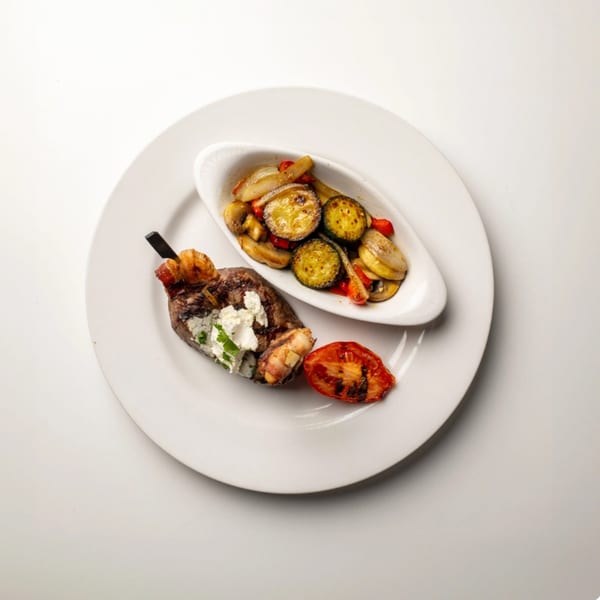 Bacon-wrapped steak with goat cheese, grilled tomato, and a side of roasted vegetables.